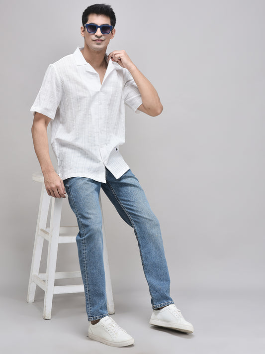 Man sitting on white stool wearing white camp collar shirt and blue jeans – with sunglasses