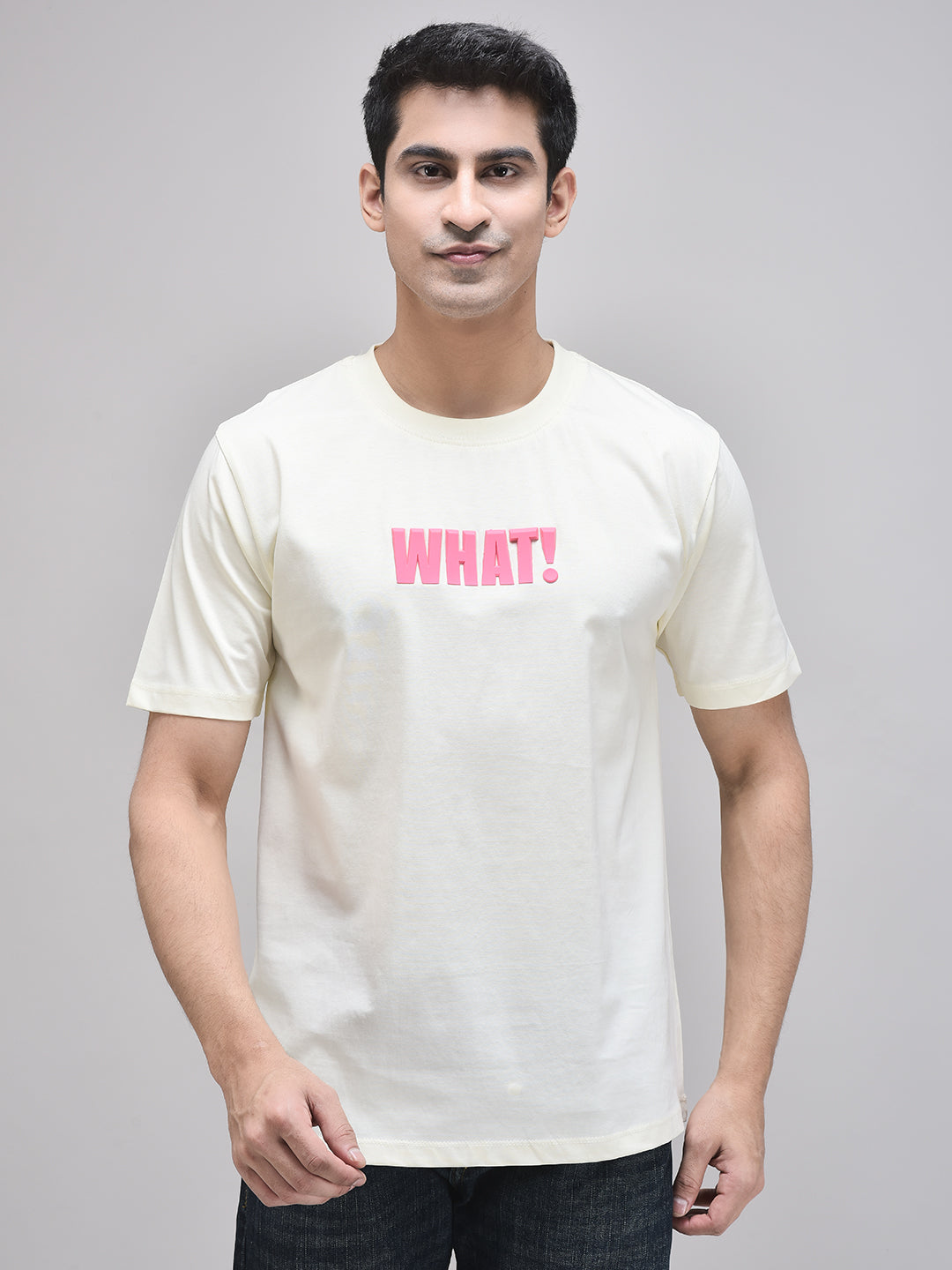 A male model wearing a beige oversized graphic t-shirt with the text 'WHAT!' printed in pink on the chest.