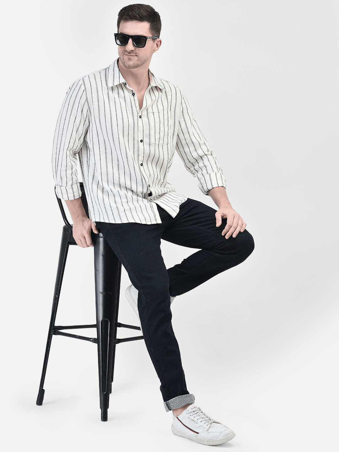A male model sitting on a stool wearing sunglasses, a striped shirt, and black trousers.