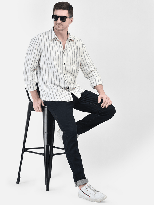 A male model sitting on a stool wearing sunglasses, a striped shirt, and black trousers.