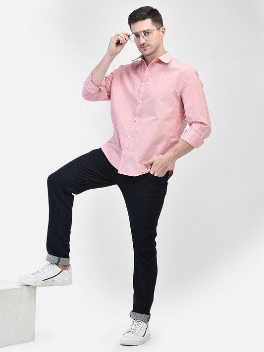 A male model wearing a pink chambray shirt and black trousers, standing on a platform, with a white and red sneaker visible in the foreground.