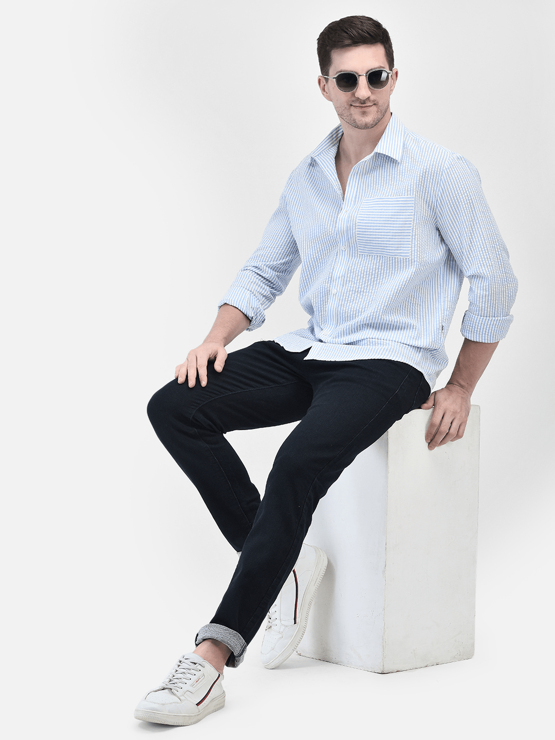 A male model wearing a blue seersucker shirt with long sleeves, paired with black trousers and sunglasses, sitting on a white stool.