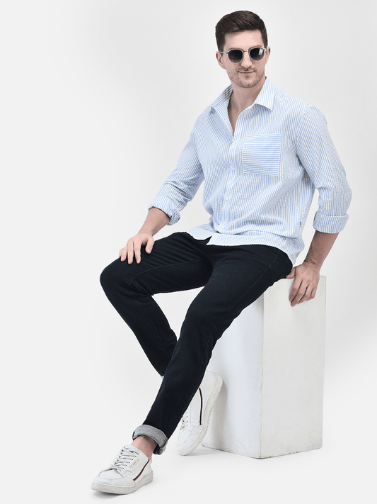 A male model wearing a blue seersucker shirt with long sleeves, paired with black trousers and sunglasses, sitting on a white stool.