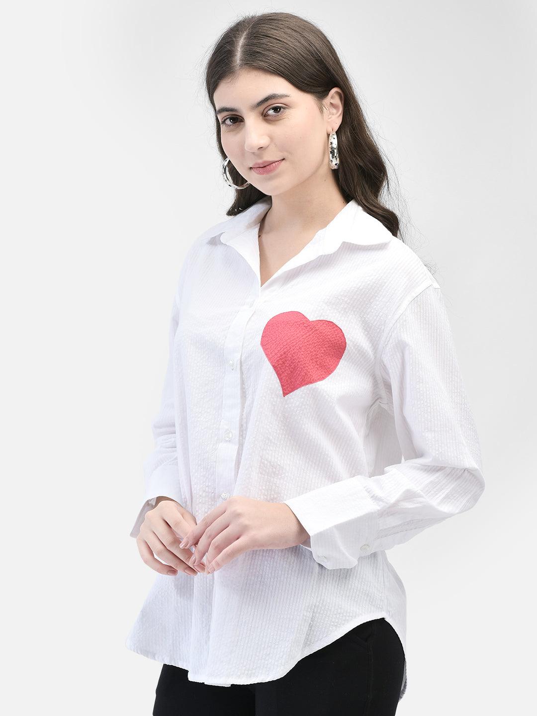 A woman wearing a white, short-sleeved, oversized shirt with a heart-shaped applique on the front, paired with black pants.