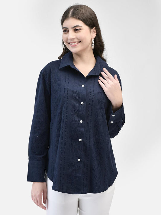 A woman smiling, wearing a navy blue, long-sleeved, button-up seersucker shirt with a collar, paired with white pants.