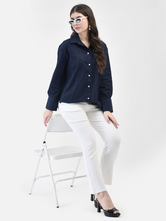 Woman seated wearing blue textured button-down shirt and white pants, stylish pose