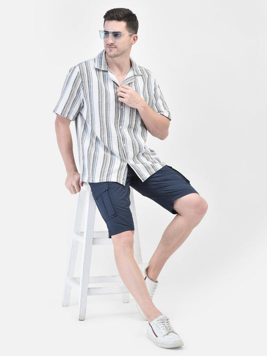 A male model wearing a white striped shirt and blue shorts, sitting on a stool.