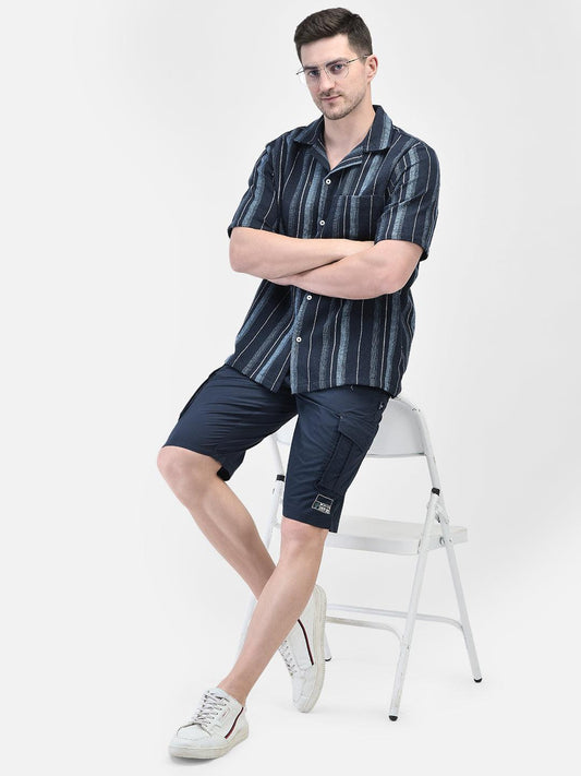 A male model wearing a navy blue and white striped shirt with short sleeves, sitting on a white chair against a white background.